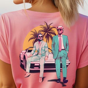 May include: Pink t-shirt featuring a graphic of two men in suits, sunglasses, and a car, with palm trees and a sunset background. The illustration has a retro, stylized design.