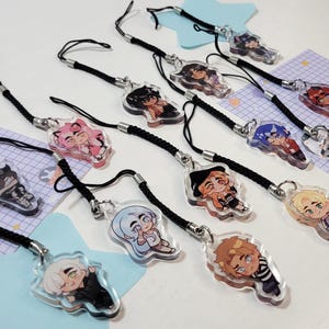 May include: A collection of 12 colorful character phone charms with black string lanyards. The charms are made of clear acrylic and feature various cartoon characters with different expressions and poses.