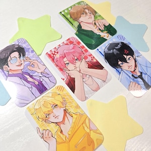 May include: Five rectangular trading cards featuring anime-style illustrations of characters with distinct hair colors including brown, pink, black, and yellow, arranged among several star-shaped paper cutouts in light green, blue, and yellow.