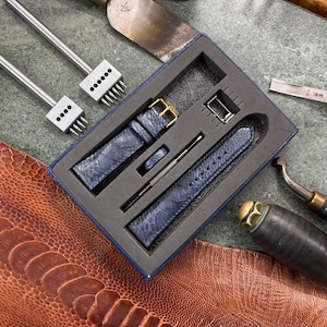 May include: A blue leather watch strap set with a gold buckle, presented in a box with leatherworking tools. The set includes a strap, buckle, and other accessories. The tools include punches and a leather cutting tool.