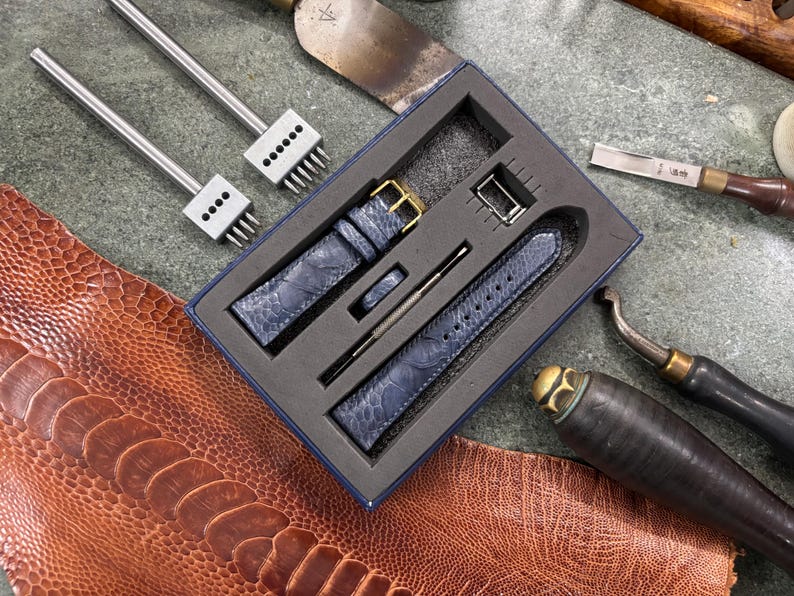 May include: A blue leather watch strap set with a gold buckle, presented in a box with leatherworking tools. The set includes a strap, buckle, and other accessories. The tools include punches and a leather cutting tool.