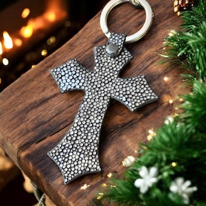 Op de afbeelding: Een zilverkleurige kruisvormige sleutelhanger met een gestructureerd, gestippeld patroon. De sleutelhanger heeft een metalen ring voor het bevestigen van sleutels. Het kruis ligt op een houten oppervlak, met kerstversiering op de achtergrond.