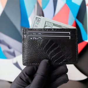 Puede incluir: Un tarjetero de cuero negro con superficie texturizada, que contiene varias tarjetas y un billete de 100 dólares. El tarjetero es sostenido por una mano enguantada, con un fondo abstracto colorido.