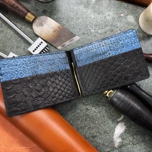 Puede incluir: Cartera de cuero abierta con un patrón de piel de serpiente azul y negro. La cartera tiene un clip de dinero dorado y varias ranuras para tarjetas. Herramientas de cuero en el fondo.