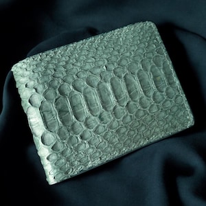 Puede incluir: Una cartera rectangular con un patrón texturizado en forma de escamas en tonos grises y verdes. La cartera está hecha de un material que se asemeja a la piel de serpiente o reptil. La cartera está cerrada y descansa sobre una tela azul oscuro.