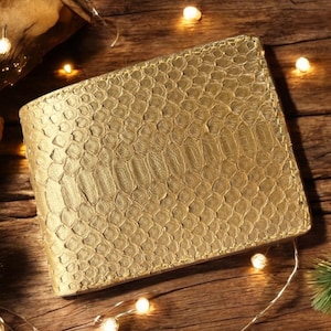 May include: A gold-colored wallet with a textured, snake-skin pattern. The wallet is rectangular and appears to be made of leather. The background is a wooden surface with small lights and greenery.