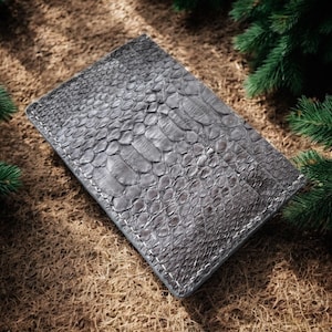 May include: A grey rectangular wallet with a snakeskin pattern. The wallet is made of a textured material and has white stitching around the edges. The wallet is on a brown surface with green pine branches in the background.