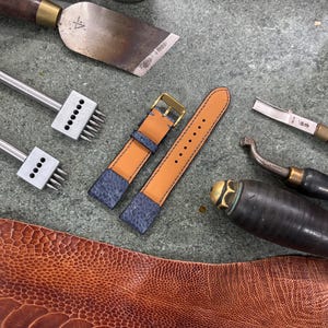 May include: A watch strap with a blue and tan leather design, alongside leatherworking tools. The strap has a gold buckle and is surrounded by various tools, including punches, a knife, and a leather chisel. The leather has a textured pattern.