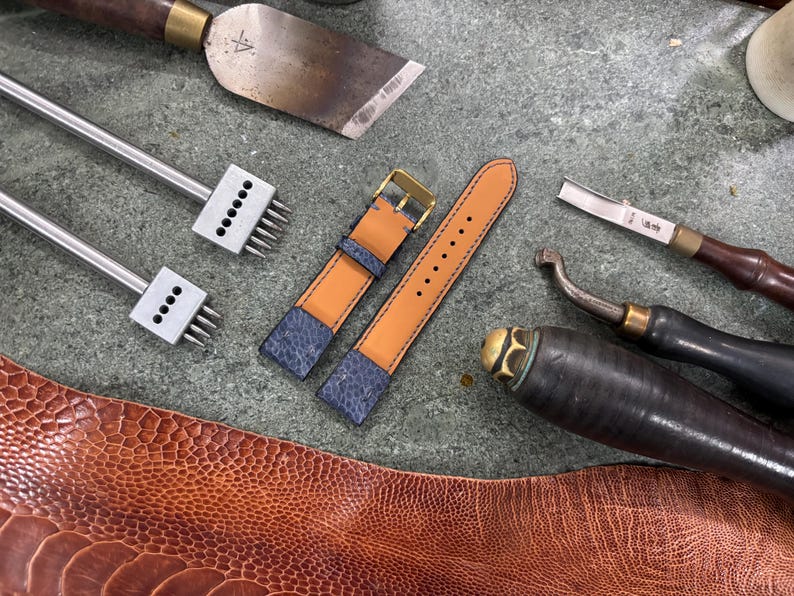 May include: A watch strap with a blue and tan leather design, alongside leatherworking tools. The strap has a gold buckle and is surrounded by various tools, including punches, a knife, and a leather chisel. The leather has a textured pattern.