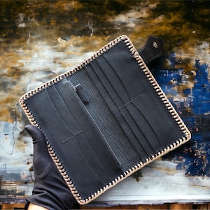 May include: A black leather long wallet featuring a textured surface, multiple card slots, and a central zippered compartment. The edges are reinforced with a thick, light-coloured whipstitch lacing that creates a distinct border around the rectangular frame.