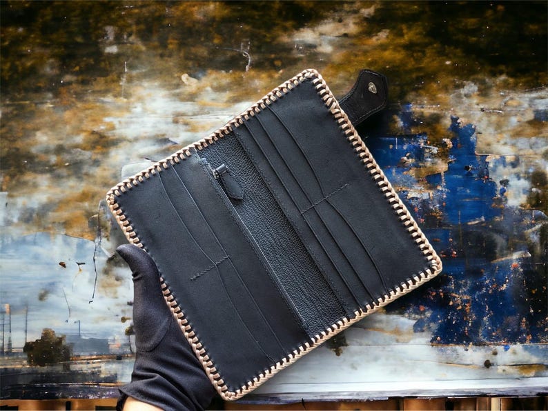 May include: A black leather long wallet featuring a textured surface, multiple card slots, and a central zippered compartment. The edges are reinforced with a thick, light-coloured whipstitch lacing that creates a distinct border around the rectangular frame.