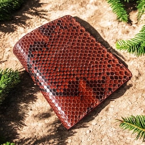 May include: A brown leather wallet with a snakeskin pattern. The wallet is rectangular and has a textured surface. The wallet is surrounded by green pine branches.