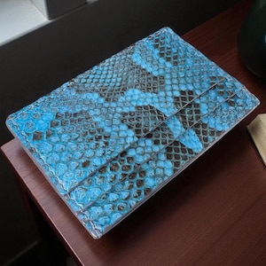 Può includere: Un portafoglio rettangolare con un motivo a pelle di serpente blu. Il portafoglio è realizzato in un materiale strutturato e ha un design a due pieghe. Il portafoglio è appoggiato su una superficie marrone scuro.