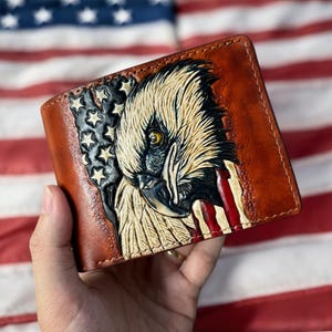 May include: Brown leather wallet with a carved eagle head design and the American flag. The bi-fold wallet is held against a backdrop of the American flag, showcasing its patriotic theme.