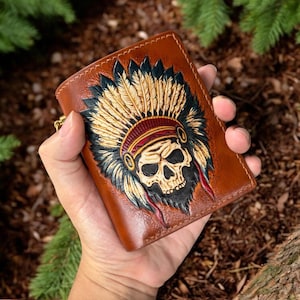Puede incluir: Una cartera de cuero marrón con un diseño grabado detallado de una calavera con un tocado de plumas. La ilustración incluye sombras y reflejos complejos en negro, rojo y beige, mostrando un acabado texturizado en la superficie.