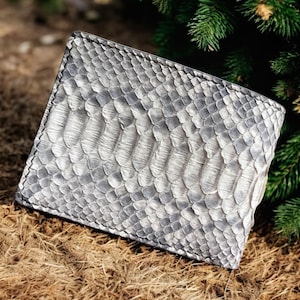 Può includere: Un portafoglio grigio e bianco con texture a pelle di serpente. Il portafoglio rettangolare sembra essere in pelle. Poggia su erba secca, con aghi di pino verdi sullo sfondo. La texture del portafoglio è molto dettagliata.