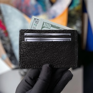 以下が含まれることがあります： 黒い革製のカードホルダー。表面はテクスチャ加工が施されており、100ドル札と2枚のカードが入っています。手袋をはめた手で持っており、コンパクトなデザインと、必需品を持ち運ぶ機能性を強調しています。