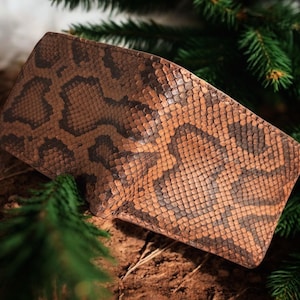 May include: A brown and tan leather wallet with a snakeskin pattern. The wallet is rectangular and appears to be made of genuine leather. The wallet is resting on a bed of dirt and surrounded by green pine needles.