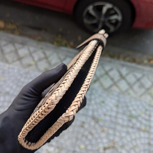May include: A bi-fold wallet featuring a black textured leather interior and exterior panels, accented by a light tan braided leather edge trim that runs along the entire perimeter of the wallet, secured with a matching braided leather tab closure.