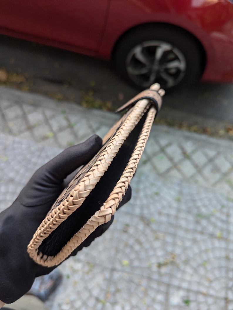 May include: A bi-fold wallet featuring a black textured leather interior and exterior panels, accented by a light tan braided leather edge trim that runs along the entire perimeter of the wallet, secured with a matching braided leather tab closure.