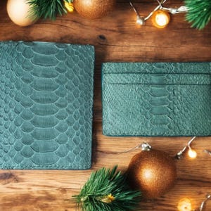 May include: A teal-colored passport holder and a matching rectangular card holder, both featuring a textured snakeskin pattern. The items are displayed side-by-side, showcasing a consistent scale-like surface design in a deep, muted green hue.