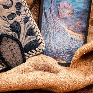 May include: A bi-fold wallet featuring intricate hand-tooled leather with a prominent skull and floral motif in tan and black. The edges are finished with a braided leather lacing, and a textured, woven-pattern tab secures the closure.