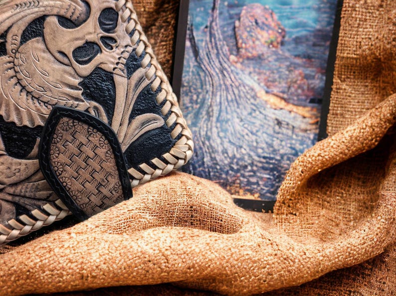 May include: A bi-fold wallet featuring intricate hand-tooled leather with a prominent skull and floral motif in tan and black. The edges are finished with a braided leather lacing, and a textured, woven-pattern tab secures the closure.