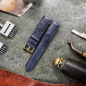 May include: A close-up of a watch strap made of blue leather with a gold buckle, alongside leatherworking tools. The tools include a blade, punches, and a curved knife, all arranged on a textured surface. The strap has a textured pattern.
