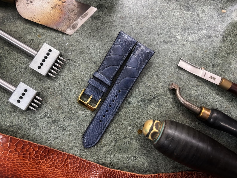 May include: A close-up of a watch strap made of blue leather with a gold buckle, alongside leatherworking tools. The tools include a blade, punches, and a curved knife, all arranged on a textured surface. The strap has a textured pattern.