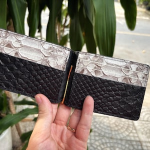 Puede incluir: Cartera de dos pliegues abierta con una sección inferior negra texturizada y una sección superior con estampado de piel de serpiente. La cartera tiene un clip para billetes dorado y varias ranuras para tarjetas. La cartera se sostiene abierta, mostrando su interior.