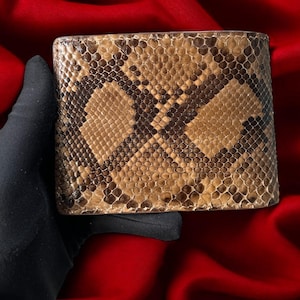 Puede incluir: Una cartera de piel de serpiente marrón y beige con un diseño de dos pliegues. La cartera tiene un patrón texturizado y se sostiene sobre un fondo rojo. La cartera parece ser nueva.