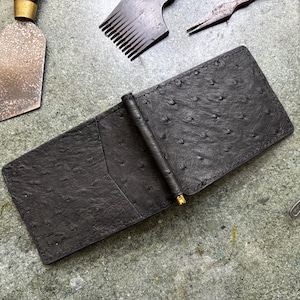 Puede incluir: Una cartera con clip para billetes de cuero negro con textura de avestruz y un mecanismo de clip metálico cilíndrico central. La cartera tiene forma rectangular con una ranura para tarjetas visible en el lado izquierdo y una textura punteada.