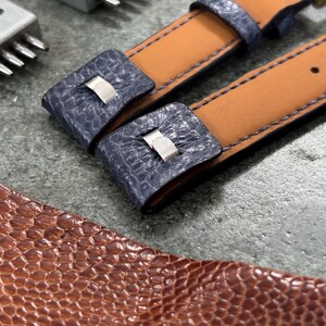 May include: Close-up of watch straps with blue textured leather accents and tan leather backing. The straps have silver-colored metal clasps and are next to a brown leather piece and a metal tool.