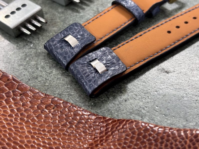 May include: Close-up of watch straps with blue textured leather accents and tan leather backing. The straps have silver-colored metal clasps and are next to a brown leather piece and a metal tool.