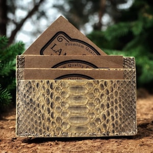 Könnte beinhalten: Ein hellbrauner Visitenkartenhalter mit Schlangenmuster. Er enthält mehrere braune Visitenkarten mit dem Text "LA" und "Premium Quality". Der Hintergrund ist verschwommen mit grünem Laub.