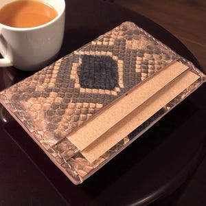 Może przedstawiać: Portfel z wzorem skóry węża z czarnym kwadratowym wzorem jest otwarty, odsłaniając dwie karty w kolorze beżowym. Biały kubek wypełniony herbatą stoi po lewej stronie na ciemnobrązowym stole. Widoczna jest faktura portfela.