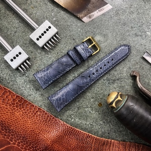 May include: A close-up shot of leather crafting tools and materials. Two blue watch straps with gold buckles are next to various metal tools, including punches and a leather knife. A piece of brown leather is in the lower left corner.
