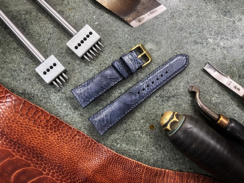 May include: A close-up shot of leather crafting tools and materials. Two blue watch straps with gold buckles are next to various metal tools, including punches and a leather knife. A piece of brown leather is in the lower left corner.