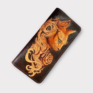 Puede incluir: Cartera de cuero marrón oscuro con un diseño en relieve detallado de una cabeza de gato y elementos florales. El gato es naranja con ojos verdes, y la cartera tiene un borde marrón oscuro. La cartera es rectangular.