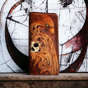 Puede incluir: Una cartera de cuero marrón con un diseño en relieve que presenta un águila y una calavera. El águila tiene plumas detalladas y una expresión feroz. La cartera es rectangular y parece estar cerrada.