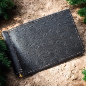 Peut inclure: Un porte-billets noir en cuir avec un motif autruche texturé. Le portefeuille a un clip doré et est cousu avec du fil marron. Le fond est une surface brun clair avec des branches de pin vertes.