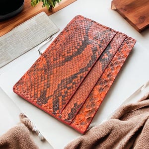 May include: A rectangular cardholder with an orange and brown snakeskin pattern. The cardholder features multiple slots and a red border. It is displayed on a white surface, alongside other decorative items.