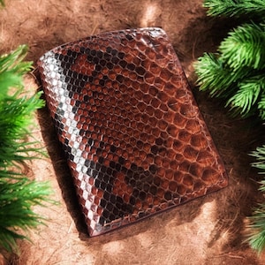 May include: A brown leather wallet with a snakeskin pattern. The wallet is rectangular and has a textured surface. The wallet is surrounded by green pine needles.