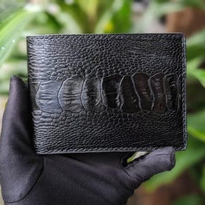 Puede incluir: Una cartera de cuero negro con una superficie texturizada y un patrón similar al de un cocodrilo. La cartera se sostiene en una mano enguantada sobre un fondo verde borroso. La cartera parece nueva.