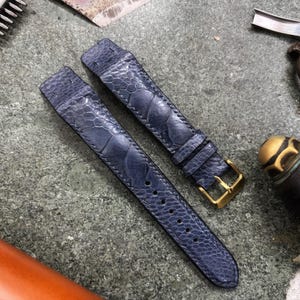 May include: Two navy blue watch straps with a textured pattern, featuring gold-colored buckles. The straps are laid out on a textured gray surface, alongside leatherworking tools, including a knife and a cylindrical tool.