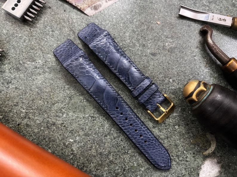 May include: Two navy blue watch straps with a textured pattern, featuring gold-colored buckles. The straps are laid out on a textured gray surface, alongside leatherworking tools, including a knife and a cylindrical tool.