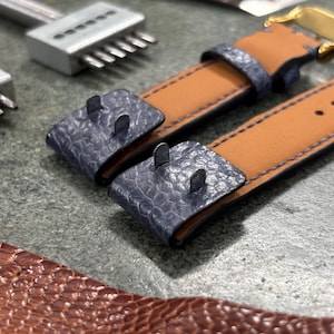 May include: Close-up of a watch strap with a tan leather interior and a blue textured exterior. The strap has a gold buckle and is next to leatherworking tools. The strap is approximately 1 inch wide.