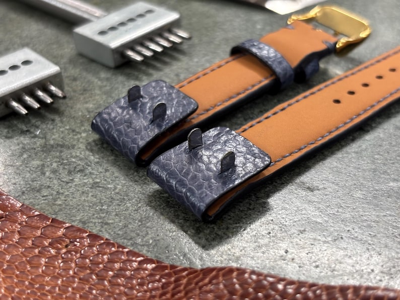 May include: Close-up of a watch strap with a tan leather interior and a blue textured exterior. The strap has a gold buckle and is next to leatherworking tools. The strap is approximately 1 inch wide.