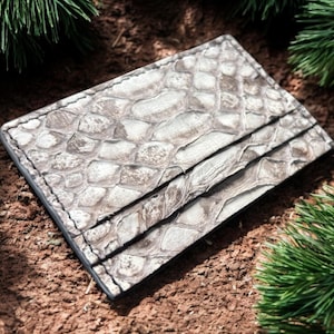 Peut inclure: Un porte-cartes gris clair avec un motif texturé semblable à celui d'un reptile. Le portefeuille comporte plusieurs fentes pour cartes et est fabriqué dans un matériau durable. Le porte-cartes est rectangulaire et semble neuf.