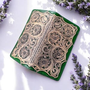 Puede incluir: Una cartera rectangular de cuero verde con un panel central que presenta intrincados patrones florales grabados a mano y una textura de tejido de cesta. El diseño en cuero natural beige contrasta con el borde verde vivo.